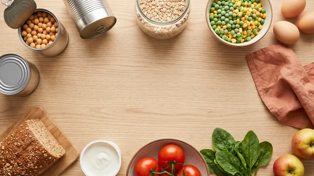 Everyday staples on a wooden table: canned chickpeas, frozen peas and corn, oats, yogurt, whole-grain bread, tomatoes, spinach and eggs
