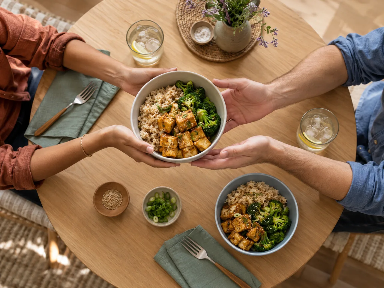 Overhead view of hands sharing bowls of brown rice, broccoli and baked tofu at a round table